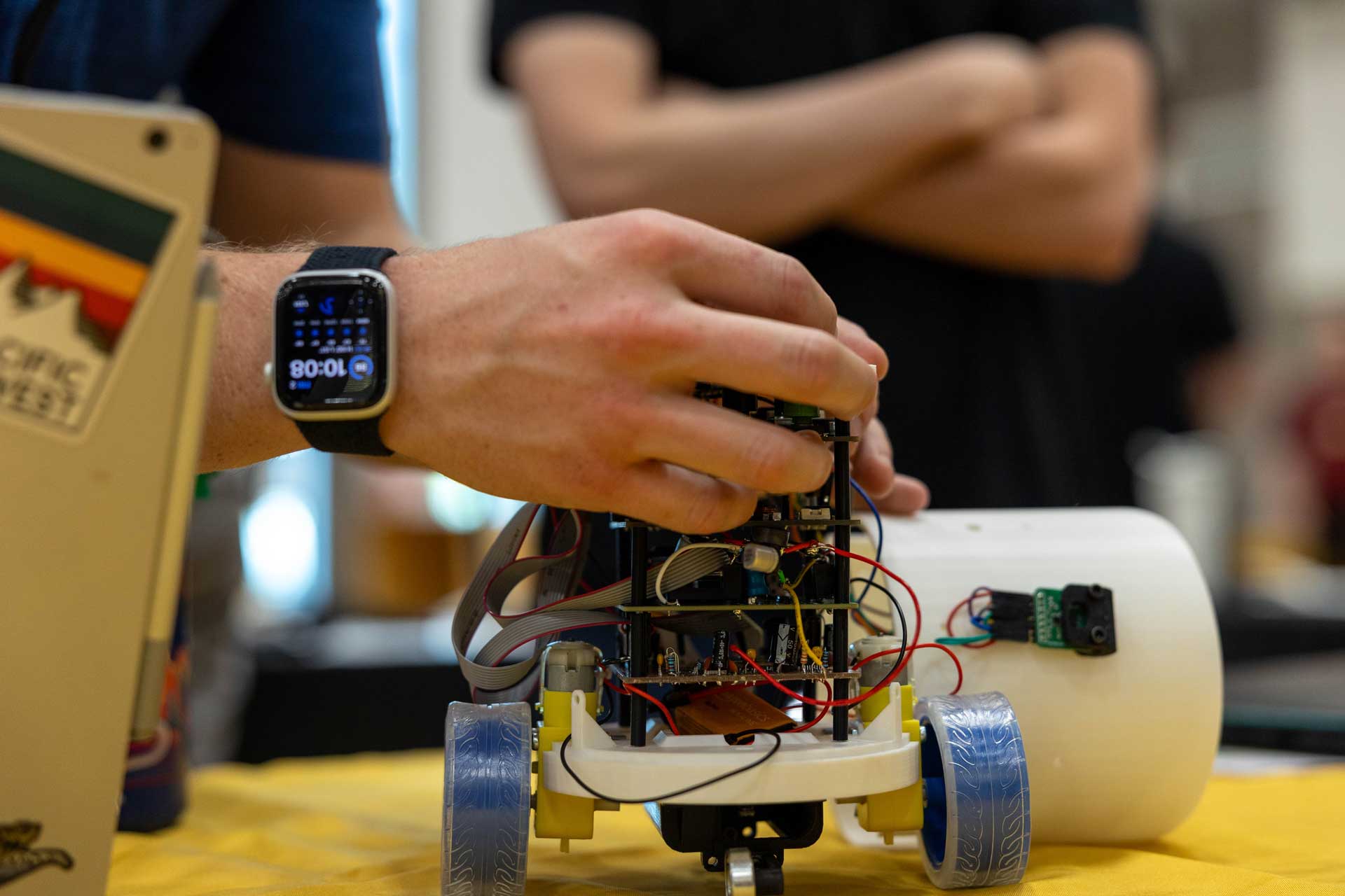 A closeup shot of a small wheeled robot being worked on by a person wearing a smartwatch.